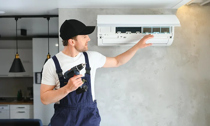 A technician with a drill inspecting an open mini-split AC unit mounted on a wall