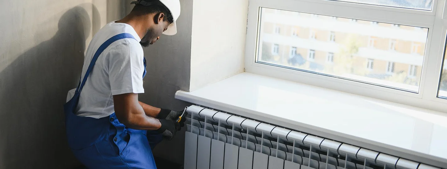 A technician in a hard hat installing a radiator beneath a window